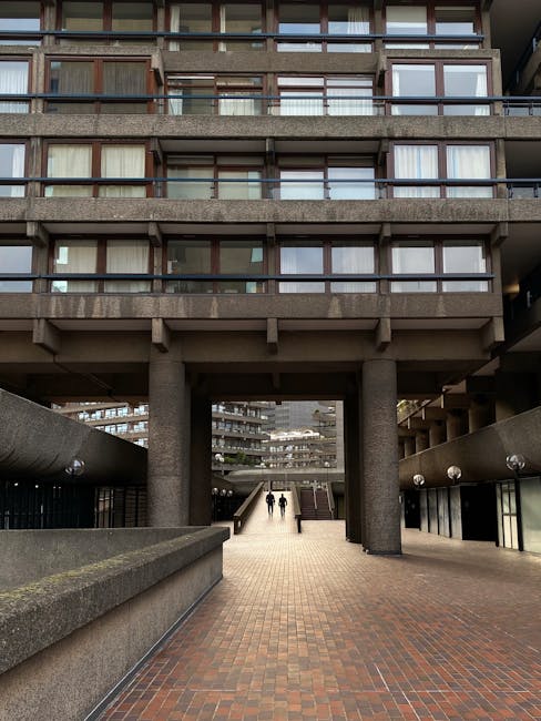 A multi-storey residential building with large windows and balconies, constructed from concrete and glass, seen from a ground-level pedestrian walkway beneath the structure. The pathway is paved with red bricks and lined with low concrete walls and cylindrical pillars supporting the upper floors. Two people are walking along the pathway towards the building, with some natural daylight illuminating the scene. This environment exemplifies urban residential architecture often encountered during home relocations or moving logistics, where clear access points for furniture transport and loading are visible. Man With a Van Barbican specializes in house removals within such settings, providing professional packing and moving services aligned with the detailed logistics of furniture transport and home relocation work.