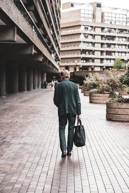 A man with curly hair wearing a dark green blazer, navy trousers, and black shoes is walking away from the camera along a paved pathway at an urban location with modern multi-storey residential buildings in the background. He is carrying a large black reusable shopping bag in his right hand. To the left, there is a building with a dark, textured facade supported by concrete columns, creating a covered walkway area. On the right, there are large, round concrete planters with greenery and bushes, and more pedestrians are visible further along the pathway. The scene is well-lit with natural daylight, and this setting appears to be part of a city environment suitable for house removals or home relocation activities. Man With a Van Barbican offers moving services in such urban areas, assisting clients with furniture transport and packing and moving logistics.