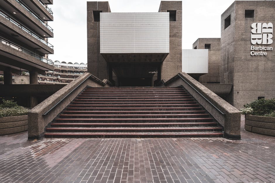 Photograph of a wide outdoor staircase leading up to the entrance of the Barbican Centre in the City of London, with brick steps and concrete railings on either side. The steps appear wet, indicating recent rain. To the right of the staircase, the large concrete building features a prominent sign reading 'Barbican Centre' with the Barbican logo above it. On the left side, residential or office buildings with balconies are visible. The area surrounding the stairs is paved with bricks, and there are planters with greenery at the base of the staircase. The scene is captured in natural daylight under an overcast sky, and the environment suggests preparation for a home relocation or furniture transport, with [COMPANY_NAME] likely involved in the moving process through their removals services.