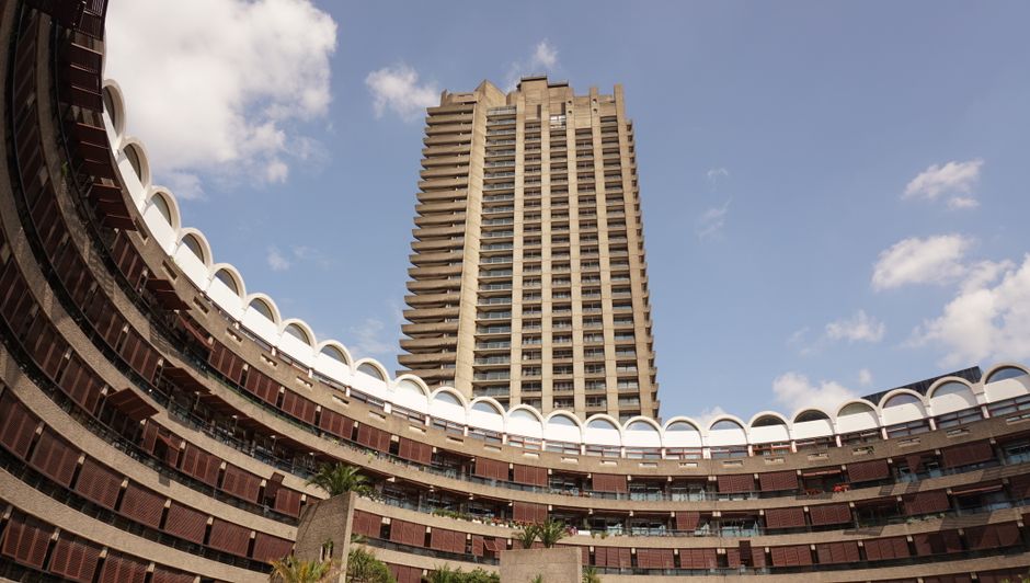 A wide-angle view of a high-rise building set against a blue sky with scattered clouds, surrounded by a circular, multi-level residential structure with curved balconies and wooden shutters. The image is taken from the ground level, looking upward, capturing part of a paved area with potted plants and trees in the foreground. Inside the curved lower balcony, several cardboard boxes, wrapped furniture, and packing materials such as bubble wrap and plastic sheeting are visible, indicating an ongoing home relocation or furniture transport process. A man from Man With a Van Barbican is seen lifting a large box onto a trolley near the entrance to facilitate smooth loading, with a van parked nearby for loading and unloading furniture and household items. The scene illustrates the logistics involved in a professional removals service focusing on packing, furniture transport, and loading activities at a residential property in the City of London, supporting the context of parking permits and city-specific moving regulations.