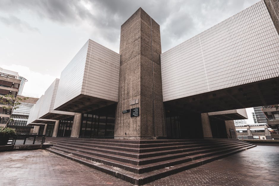 A modern, institutional-style building with exterior steps leading up to an entrance, featuring a central concrete column flanked by large, white tiled sections that extend outward on either side, creating a cantilevered effect. The steps are made of dark brick and are arranged in a symmetrical, tiered fashion, leading up to glass doors that form the building’s entryway. Surrounding the building are neighboring structures, with a partly cloudy sky overhead providing diffuse natural light. In the foreground, there are no visible vehicles or loading equipment, though the area appears to be an accessible, open paved space suitable for moving or loading activities. This image, associated with home relocation and furniture transport, supports visual understanding of building access points, which are relevant to professional removals services such as those offered by Man With a Van Barbican, involved in packing, moving, and transport logistics.