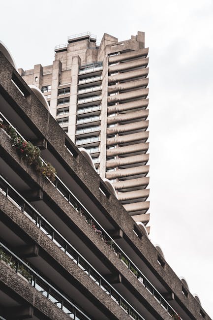 Photograph of a multi-storey residential building with long, curved balconies featuring metal railings and some with potted plants and flowers, viewed from below. In the foreground, part of a concrete block parking structure is visible, with a smooth surface and overhanging balcony edge. The sky is overcast, casting diffused light over the scene. The image reflects an urban environment suitable for house removals and moving services, with a focus on vertical living spaces often involved in home relocation processes, as carried out by Man With a Van Barbican. The perspective emphasizes the height of the building and the structural details of the balconies, illustrating the typical characteristics of city apartment blocks in the Barbican area.
