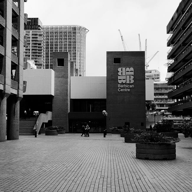 A man wearing a dark green jacket and dark jeans is walking along a paved sidewalk in an urban environment, carrying a black striped bag in his right hand. The scene features modern multi-storey residential buildings with balconies and large windows on the right side, and a raised structure supported by concrete pillars on the left. There are large planters with small trees and shrubs along the pathway, and a few other pedestrians are visible in the distance. The lighting suggests daytime with natural light illuminating the scene, capturing an active home relocation process or movement of belongings. The image is natural and professional, reflective of a city-based moving service such as Man With a Van Barbican, with a focus on logistics, packing, or transport during house removals.