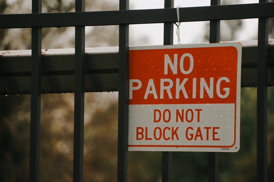 A close-up view of a rectangular safety sign mounted on a metal fence with vertical bars. The sign features a red and white colour scheme and displays the messages 'NO PARKING' in large white letters on a red background, followed by 'DO NOT BLOCK GATE' in smaller red letters on a white background. The sign is affixed with a metal wire at the top. The background appears blurred with natural outdoor lighting, indicating the fence is part of a property boundary, likely near a driveway or gate area. This image relates to house removals or moving services by highlighting restrictions that may impact loading and unloading zones during a home relocation, which Man With a Van Barbican services in the EC2, Barbican area might encounter during furniture transport and packing and moving processes.