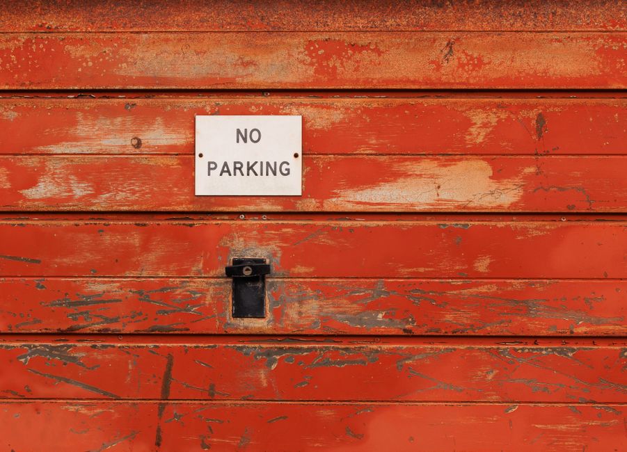 Close-up of a weathered red wooden wall with peeling paint, featuring a small white sign that reads 'NO PARKING' in black text. Below the sign, an old black lock mechanism is mounted on the wall. The surface shows signs of age and exposure to the elements. This image captures a typical urban setting where loading and unloading activities for house removals may be restricted by such signage. The focus on the wall, sign, and lock highlights the challenges faced during home relocation, especially when loading bays or parking areas are limited or regulated. Man With a Van Barbican, a company specialising in removals, often encounters such restrictions in the EC2 Barbican area, requiring careful planning for furniture transport and loading processes. The environment suggests an exterior setting, with neutral lighting emphasizing the textures of the aged wood and the signage.