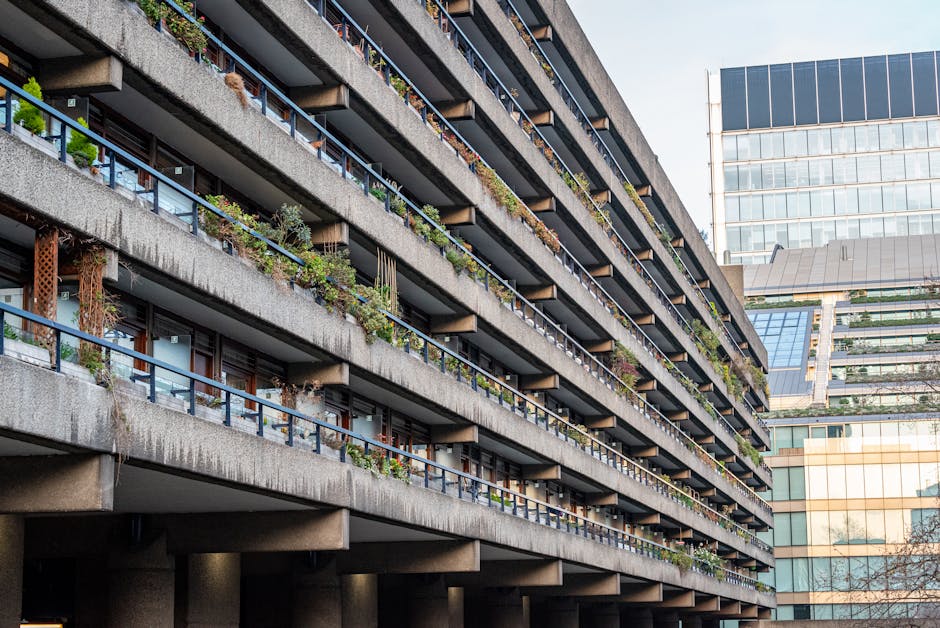 A multi-storey residential building featuring concrete balconies with blue metal railings, each adorned with potted plants and small garden decorations. The building's exterior shows signs of weathering, with some discolouration. In the foreground, part of a Man With a Van Barbican vehicle is visible, indicating a home relocation or furniture transport activity in progress. The urban environment is characterized by adjacent modern glass office buildings with reflective surfaces. The scene is captured during daylight hours with clear skies, and the overall setting depicts the logistical aspect of moving services in a complex, high-access residential area. This image is relevant to house removals and the logistics involved in narrow access moving scenarios, highlighting the importance of professional packing, loading, and transport planning.