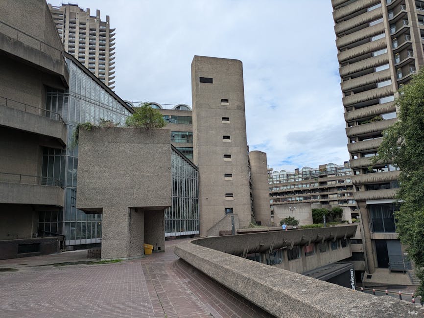 A wide view of a modern urban residential area featuring Brutalist-style concrete buildings with multiple levels and rounded balconies, set against a partly cloudy sky. In the foreground, there is a paved open space with a low, sloped concrete retaining wall. To the left, a section of a building with large glass windows and a balcony is visible, with a small yellow bucket placed near the entrance. In the middle ground, a narrow alleyway leads to the rear entrance of the building, which has a small door and a person in casual clothing walking nearby. Tall, vertically aligned concrete structures, characteristic of the estate, dominate the scene, with some greenery visible on ledges and rooftops. To the right, several trees frame the scene, providing contrast against the concrete architecture. This setting relates to home relocation and furniture transport within urban residential developments, as handled by Man With a Van Barbican, highlighting the logistics involved in house removals on narrow estate access points.