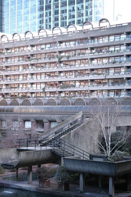 A multi-storey residential building featuring concrete balconies with blue metal railings, each adorned with potted plants and small garden decorations. The building's exterior shows signs of weathering, with some discolouration. In the foreground, part of a Man With a Van Barbican vehicle is visible, indicating a home relocation or furniture transport activity in progress. The urban environment is characterized by adjacent modern glass office buildings with reflective surfaces. The scene is captured during daylight hours with clear skies, and the overall setting depicts the logistical aspect of moving services in a complex, high-access residential area. This image is relevant to house removals and the logistics involved in narrow access moving scenarios, highlighting the importance of professional packing, loading, and transport planning.