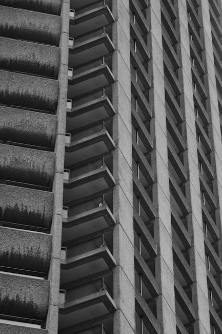 A black-and-white photograph of a high-rise residential building with multiple balconies arranged vertically on the left side of the image. The building's exterior is composed of concrete with visible texture and horizontal lines, indicating a modern structural design. The balconies have metal railings and some have visible items such as small plants or furniture, though details are indistinct due to the monochrome tone. The right side of the image shows a section of another building with a similar concrete facade and diagonal window arrangements, creating a geometric pattern. The environment appears to be an urban setting, likely within the Barbican estate area, and the photo emphasizes the height and architectural features of the residential towers. This structure provides context for discussions on house removals or moving logistics within dense city environments, supporting services like those offered by Man With a Van Barbican.