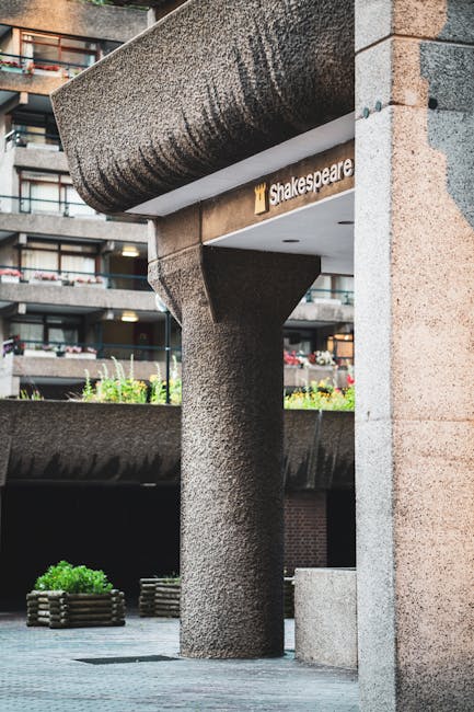 A close-up view of the exterior of a building showing a textured, rounded concrete pillar supporting a large overhanging concrete canopy. The building features a sign with the logo and name of Shakespeare, partially visible beneath the canopy. In the background, there are residential balconies with potted plants and flowers, indicating an urban residential area. The scene is well-lit with natural daylight, and the ground in front of the building includes a paved area with a low, curved planter containing greenery. This setting captures the environment where professional house removals, such as those from Man With a Van Barbican, may involve navigating narrow doorways and tight spaces during home relocation or furniture transport, emphasizing the importance of careful planning in lifting and loading furniture through confined access points.
