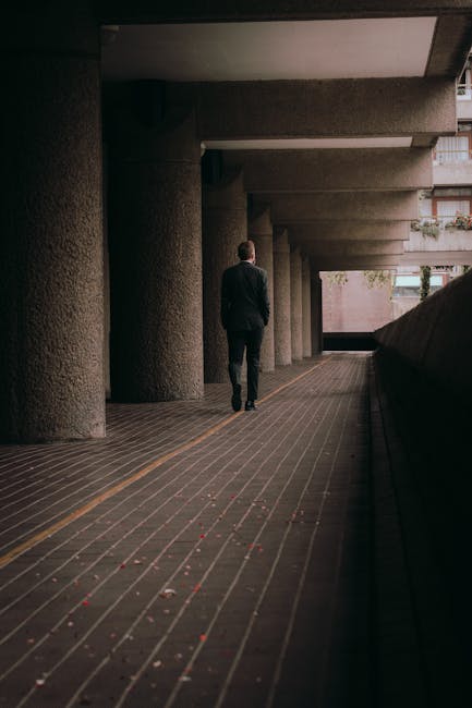 A man wearing a dark suit is walking away from the camera along an outdoor covered walkway beneath a multi-storey building structure supported by large concrete columns. The walkway features a tiled surface with a yellow line running parallel to the building's edge, and small scattered debris is visible on the ground. To the right, there is a dark vehicle parked close to the edge of the walkway, partially visible in the frame. The scene is lit by natural daylight filtering through the open sides of the corridor. This setting appears to be part of an urban environment, suitable for house removals or moving services, as it depicts the process of carrying household items through shared communal spaces before loading onto a van, consistent with professional home relocation activities provided by companies like Man With a Van Barbican.