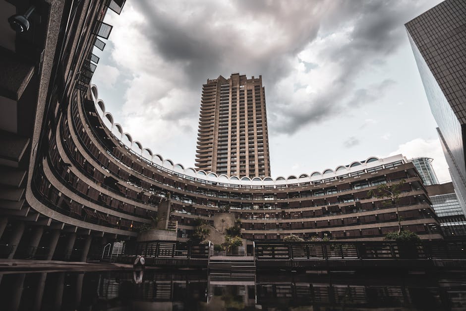 Photograph taken from ground level looking up at the curved exterior of a multi-storey building within an urban area, with a tall high-rise tower in the background under a cloudy sky. The curved building features multiple floors with open balconies bordered by white railings, and each floor has a row of dark windows. The area in front of the building includes a paved surface with a few small trees and a black metal fence. The scene captures the environment where home relocation services provided by Man With a Van Barbican might involve loading or unloading furniture and boxes from nearby vehicles, in an area characterized by modern architecture and city infrastructure.