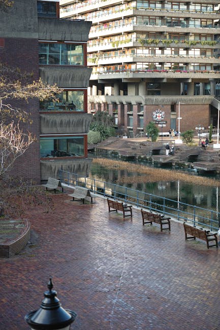 A man wearing a dark suit is walking away from the camera along an outdoor covered walkway beneath a multi-storey building structure supported by large concrete columns. The walkway features a tiled surface with a yellow line running parallel to the building's edge, and small scattered debris is visible on the ground. To the right, there is a dark vehicle parked close to the edge of the walkway, partially visible in the frame. The scene is lit by natural daylight filtering through the open sides of the corridor. This setting appears to be part of an urban environment, suitable for house removals or moving services, as it depicts the process of carrying household items through shared communal spaces before loading onto a van, consistent with professional home relocation activities provided by companies like Man With a Van Barbican.