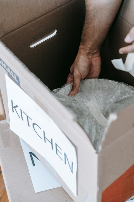 Close-up view of a person’s hand inserting an item into a cardboard box labeled 'KITCHEN' during a house removal process. The box is partially packed with air-filled plastic bubble wrap visible at the top for protective packaging. The scene is set indoors, near a carpeted floor, with a brown cardboard moving box placed on a flat surface. The background includes a section of a door or doorway, indicating an interior environment suitable for packing and moving. The image captures the careful packing activity typical of professional remova ls by Man With a Van Barbican, supporting efficient furniture transport and home relocation tasks, particularly during packing for transfers like Silk Street Moves in EC2Y.