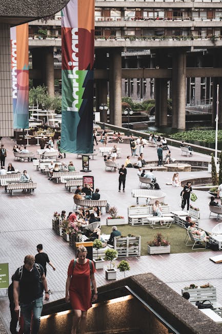 The image shows an urban outdoor seating area beneath a large bridge or overpass, with multiple people sitting on benches and at tables, some engaged in conversations or using electronic devices. The space is decorated with numerous potted plants and wooden planters, creating a casual, landscaped environment. In the foreground, a woman in a red dress and a man in a black shirt walk down stone stairs, heading toward the seating area. Behind the scene, several individuals are walking or standing, carrying shopping bags or personal items. Large vertical banners with colorful branding hang from tall poles, and the surrounding architecture includes concrete supports for the bridge above. This setting is likely part of a city development project or recreational zone, often used for community gathering or social events. The image’s context emphasizes urban mobility and public areas, which relate indirectly to the logistics of home relocation and furniture transport managed by companies like Man With a Van Barbican, providing essential delivery and packing services for house moves.