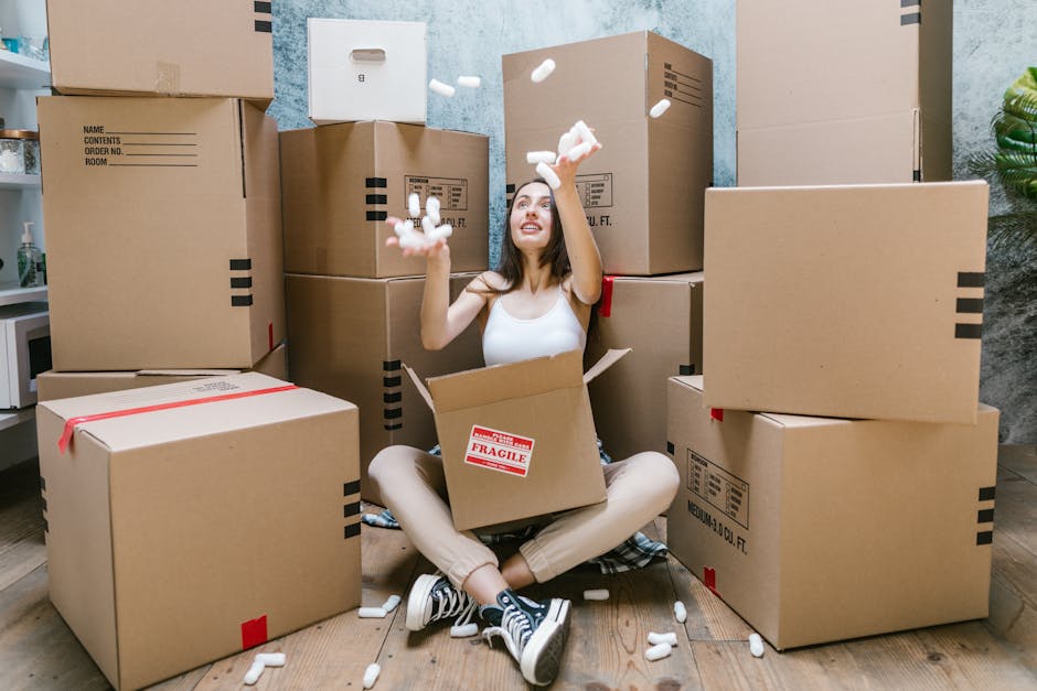 Close-up view of a person’s hand inserting an item into a cardboard box labeled 'KITCHEN' during a house removal process. The box is partially packed with air-filled plastic bubble wrap visible at the top for protective packaging. The scene is set indoors, near a carpeted floor, with a brown cardboard moving box placed on a flat surface. The background includes a section of a door or doorway, indicating an interior environment suitable for packing and moving. The image captures the careful packing activity typical of professional remova ls by Man With a Van Barbican, supporting efficient furniture transport and home relocation tasks, particularly during packing for transfers like Silk Street Moves in EC2Y.