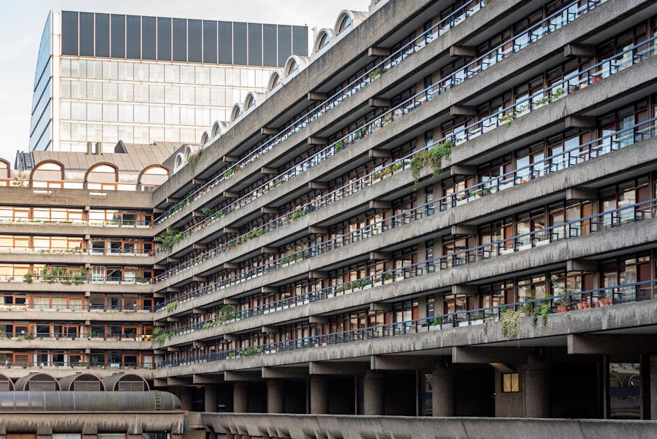 Photograph of a multi-storey residential building with exterior balconies lined with black metal railings, some with potted plants and outdoor furniture, set against a background of a modern office building with glass windows. The building's facade features exposed concrete surfaces, and the bottom level shows a parking or loading area underneath supported by large concrete columns. The scene, captured during daylight with natural lighting, illustrates urban apartment living, relevant to house removals and home relocation services provided by Man With a Van Barbican, focusing on logistics like moving furniture and boxes across multi-level properties.