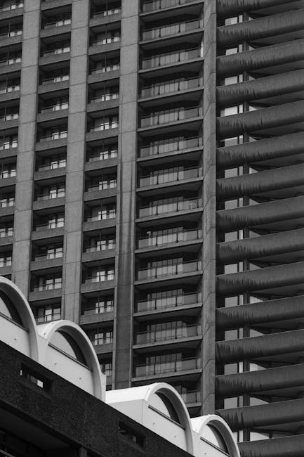 Black and white photograph showcasing the exterior of a high-rise residential building with multiple balconies and windows, alongside a modern multi-level parking structure with curved white arches on the roof. The scene appears to be in an urban area, possibly near the Barbican estate. The parking structure's concrete surface contrasts with the textured facade of the building. This image may illustrate urban housing or relocation logistics, relevant to house removals and furniture transport. The photograph's perspective emphasizes the height and architectural details of the building, with natural lighting highlighting the different materials. Man With a Van Barbican could utilize this image to illustrate the environment where home relocation or packing and moving activities take place, especially for last-minute or same-day removal services.