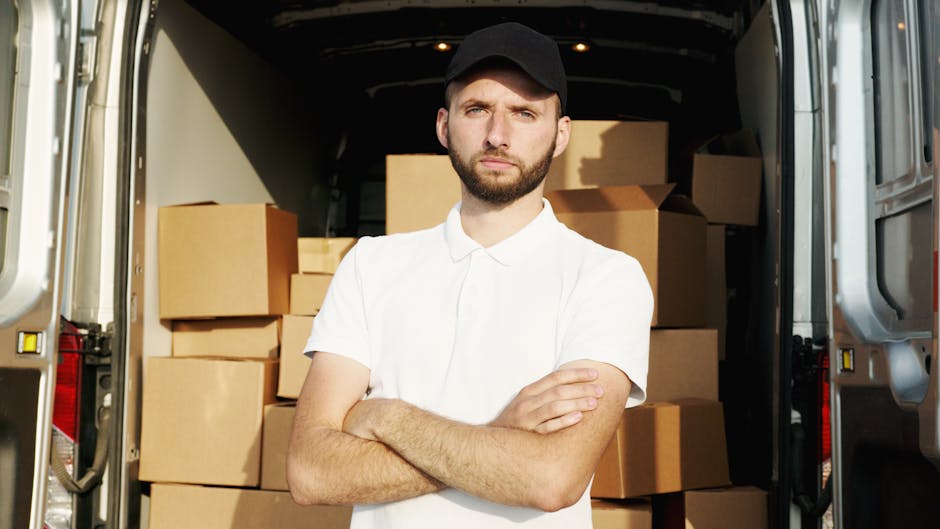 Three individuals, two women and one man, are positioned inside the back of a white moving van parked outdoors. The van's rear doors are open, revealing several packed cardboard boxes of varying sizes