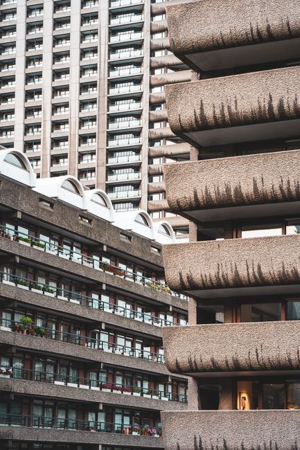 A multi-storey residential building with rows of balconies featuring glass balustrades and potted plants, situated in an urban environment with other high-rise structures visible in the background. Th